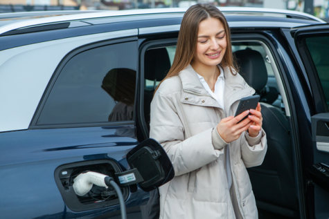Moteris įsikrauna elektromobilį ir tuo pat metu naršo telefone