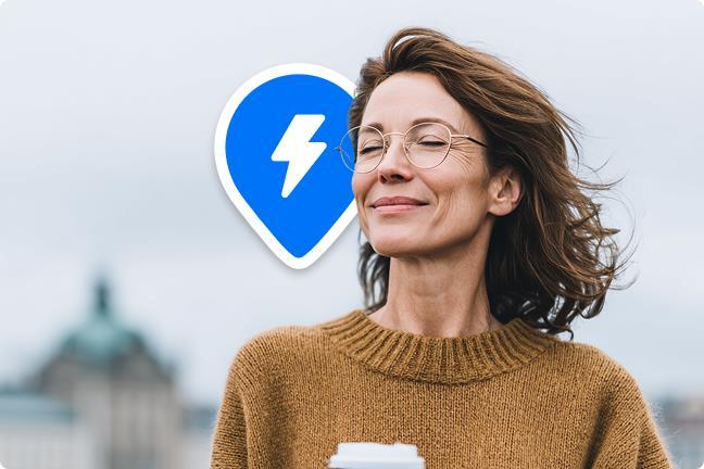 Besišypsanti moteris su kavos puodeliu rankoje, o fone Ignitis ON įkrovimo stotelės PIN ženkliukas.