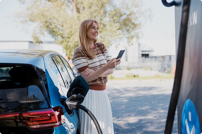 Besišypsanti moteris, atsirėmusi į elektromobilį, su mobiliu telefonu rankoje, kraunasi elektromobilį Ignitis ON įkrovimo stotelėje.