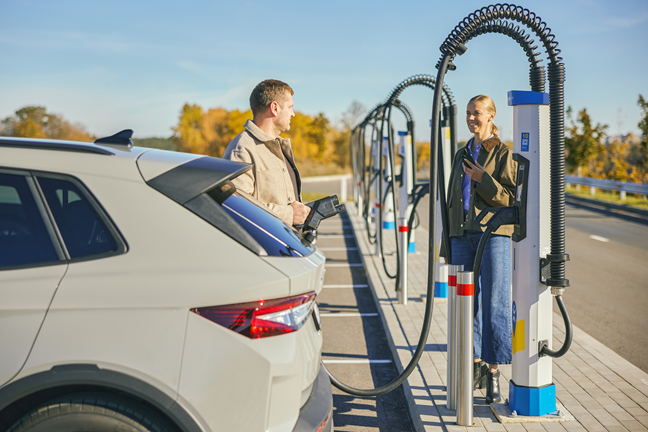 Vyras ir moteris žiūri vienas į kitą ir šypsosi. Vyras rankoje laiko įkrovimo jungtį ir ruošiasi įkrauti savo elektromobilį Ignitis ON stotelėje.