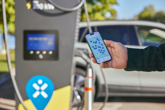 Rankoje mobilus telefonas, kurio ekrane įjungtas Ignitis ON programėlės įsikrovimo lokacijų žemėlapis. Fone stovi Ignitis ON greito įkrovimo stotelė.