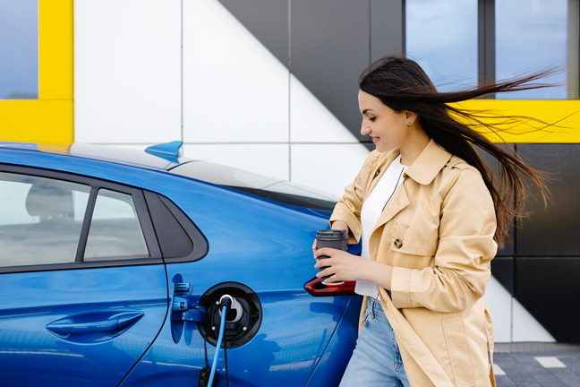 Besišypsanti moteris su kavos puodeliu rankoje įsikrauna savo elektromobilį.